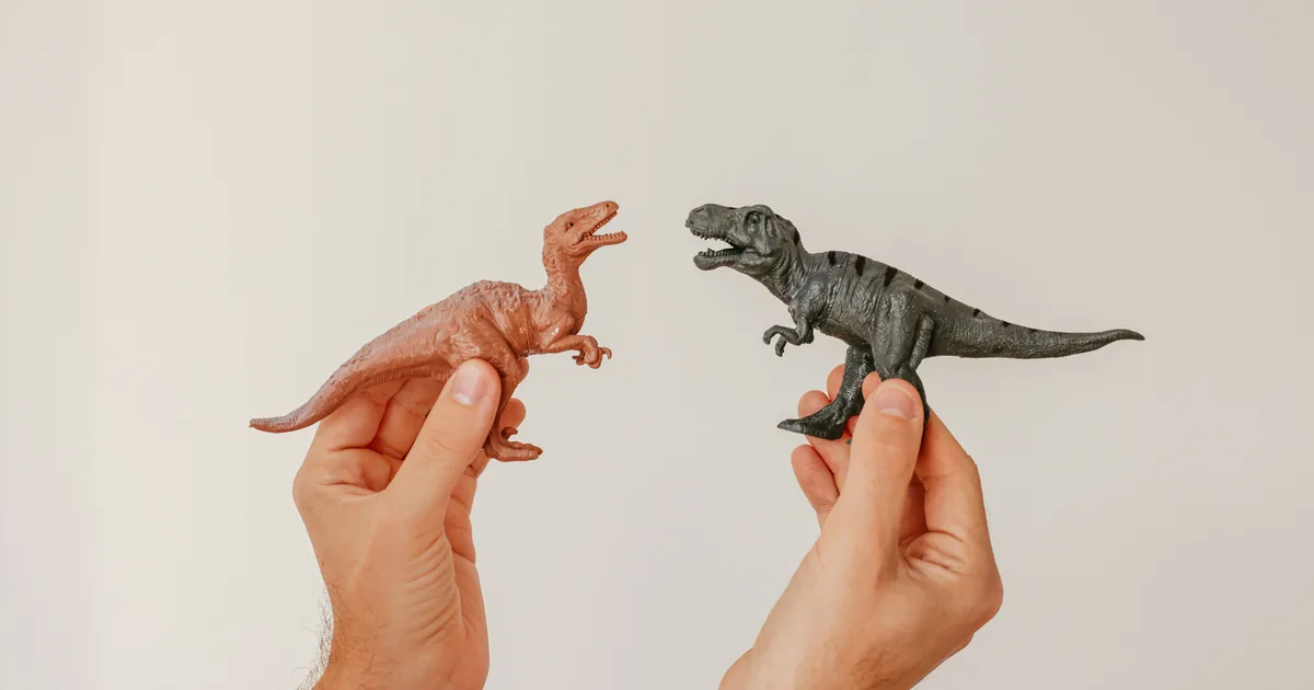 Deux mains tenant face à face deux figurines de dinosaures, l'une brune, l'autre noire, sur fond crème